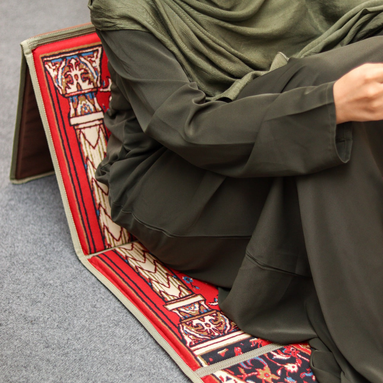 a woman in abaya sitting on ebadat prayer rug colored lava red with its backrest opened