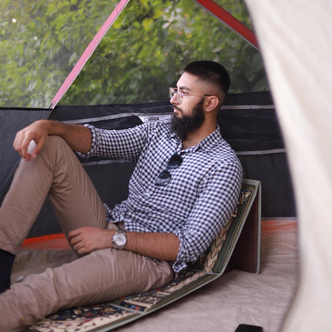 a man with glasses sitting on an earth green ebadat prayer rug with the backrest opened